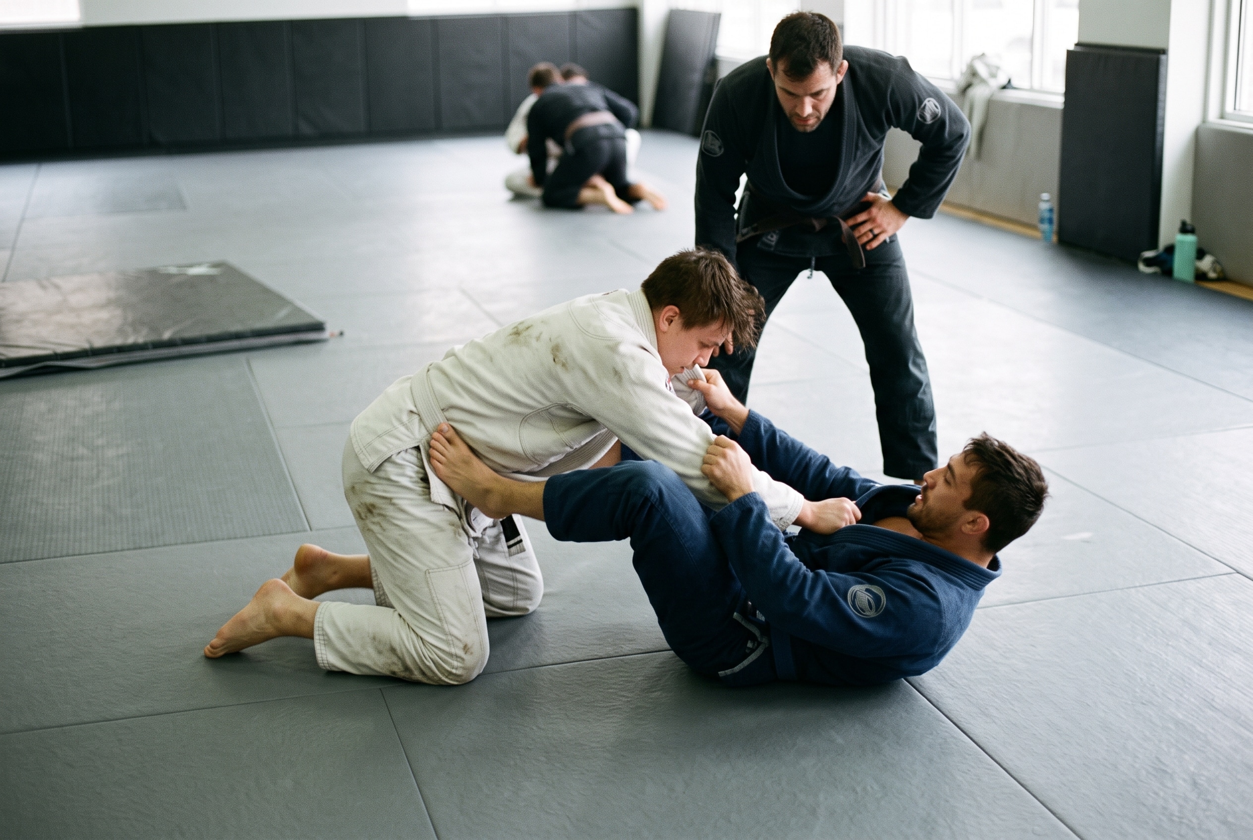 A candid, natural-light photograph capturing a close-up of practitioners at a Brazilian Jiu-Jitsu (BJJ) academy, illustrating BJJ Injuries: What Gym Owners Need to Know About Risk and Coverage and the intense physical contact that creates operational risk. In the center, two grappling students wearing traditional gis (one in white, one in dark blue) are actively rolling on grey tatami mats, with the student in the white gi on his knees attempting a guard pass while the student in blue defends from his back.