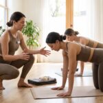 A yoga insurance cost instructor kneels beside a student holding a tabletop pose, gesturing with open hands to guide alignment and safe form in a sunlit yoga studio.